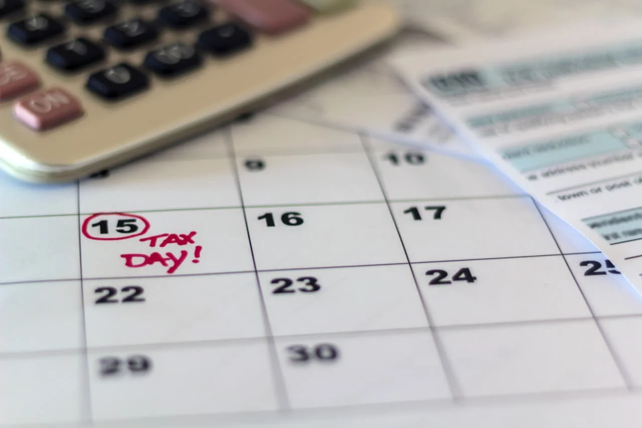 Um calendário de mesa elegante sobre uma escrivaninha de madeira, com o dia 15 de abril circulado em vermelho, ao lado de um gráfico de ações ascendente em um tablet.