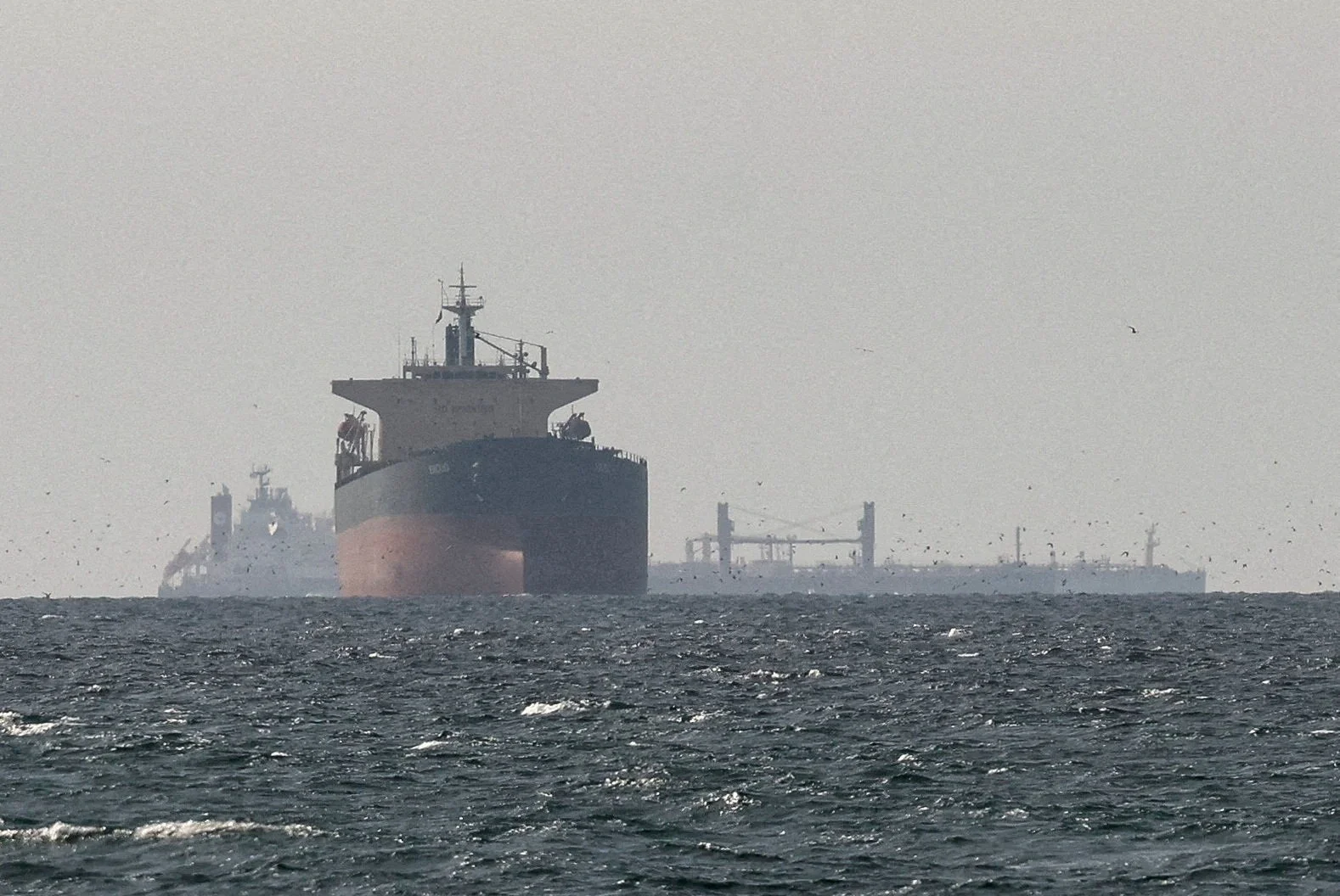 Navios-tanque navegando em formação no Estreito de Ormuz, com montanhas áridas ao fundo e um céu azul claro. A cena transmite a importância da rota marítima para o comércio global de petróleo.