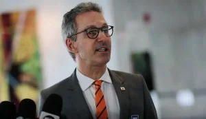 Um político brasileiro de meia-idade, com cabelos curtos e óculos, em um palanque, gesticulando para uma plateia em um evento de campanha eleitoral, com bandeiras ao fundo.