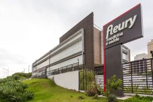 Uma mão segura um smartphone com o logotipo da Fleury (FLRY3) em um gráfico de ações, enquanto moedas brasileiras (reais) caem suavemente ao fundo, simbolizando dividendos e ganhos financeiros.