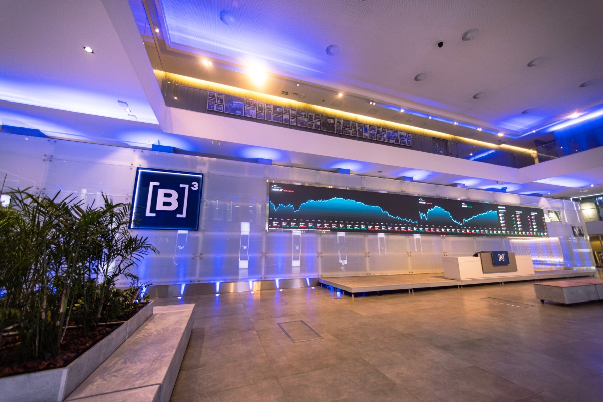 Painel digital de mercado financeiro exibindo o índice Ibovespa em alta com números verdes, ao fundo uma imagem desfocada de um terminal portuário de petróleo.