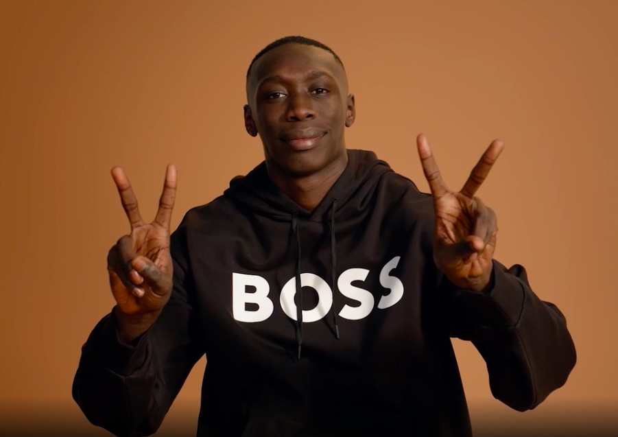 Uma pessoa jovem, de pele escura e feições expressivas, semelhante a Khaby Lame, com uma expressão de confusão ou mistério, em meio a gráficos financeiros em queda e símbolos de Wall Street ao fundo, em tons escuros e azuis.