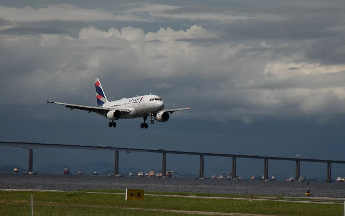 Aeronave moderna com pintura azul e vermelha decolando de uma pista de aeroporto ao entardecer, com o horizonte urbano de São Paulo ao fundo sob luz dourada.