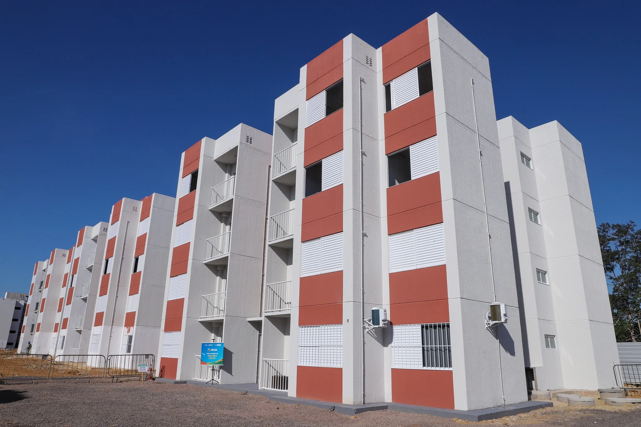 Fachada de um condomínio residencial moderno com prédios de cores claras, janelas amplas e áreas verdes ao redor, sob a luz do pôr do sol.