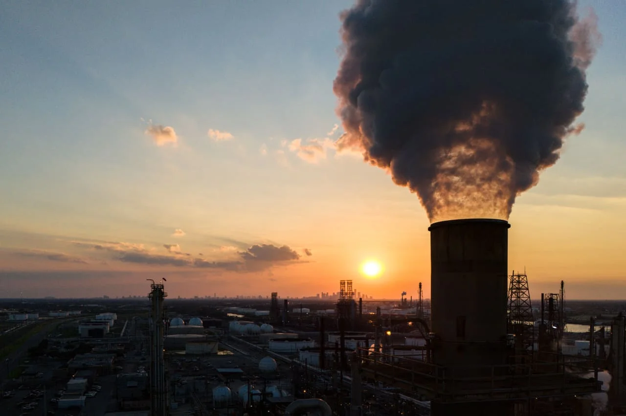 Visão cinematográfica de barris de petróleo empilhados em um porto ao entardecer, com gráficos de velas financeiras flutuando em tons de dourado e azul.