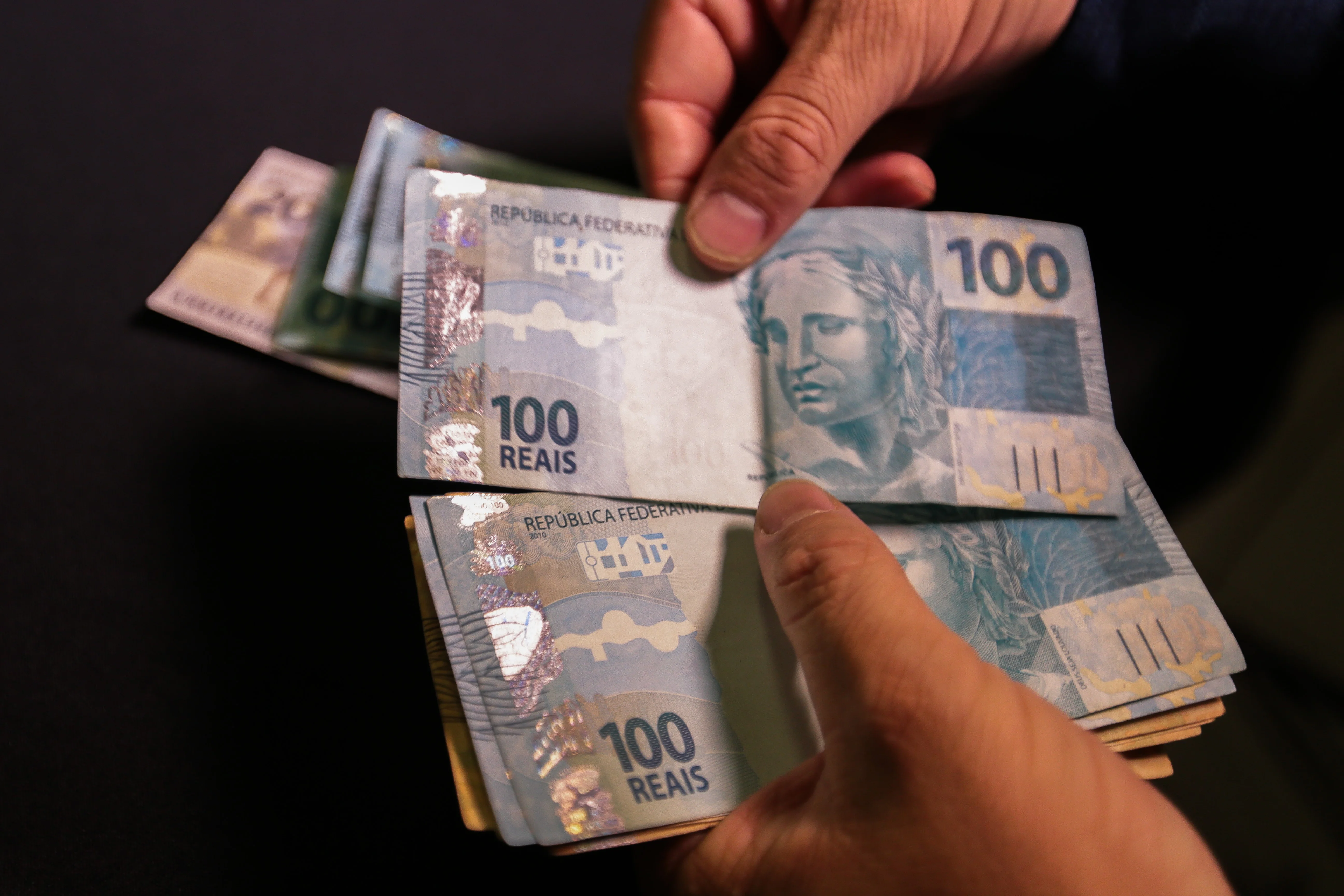 Close-up de mãos brasileiras contando notas de 100 reais sobre uma mesa de madeira, com uma calculadora e boletos ao fundo em ambiente doméstico.